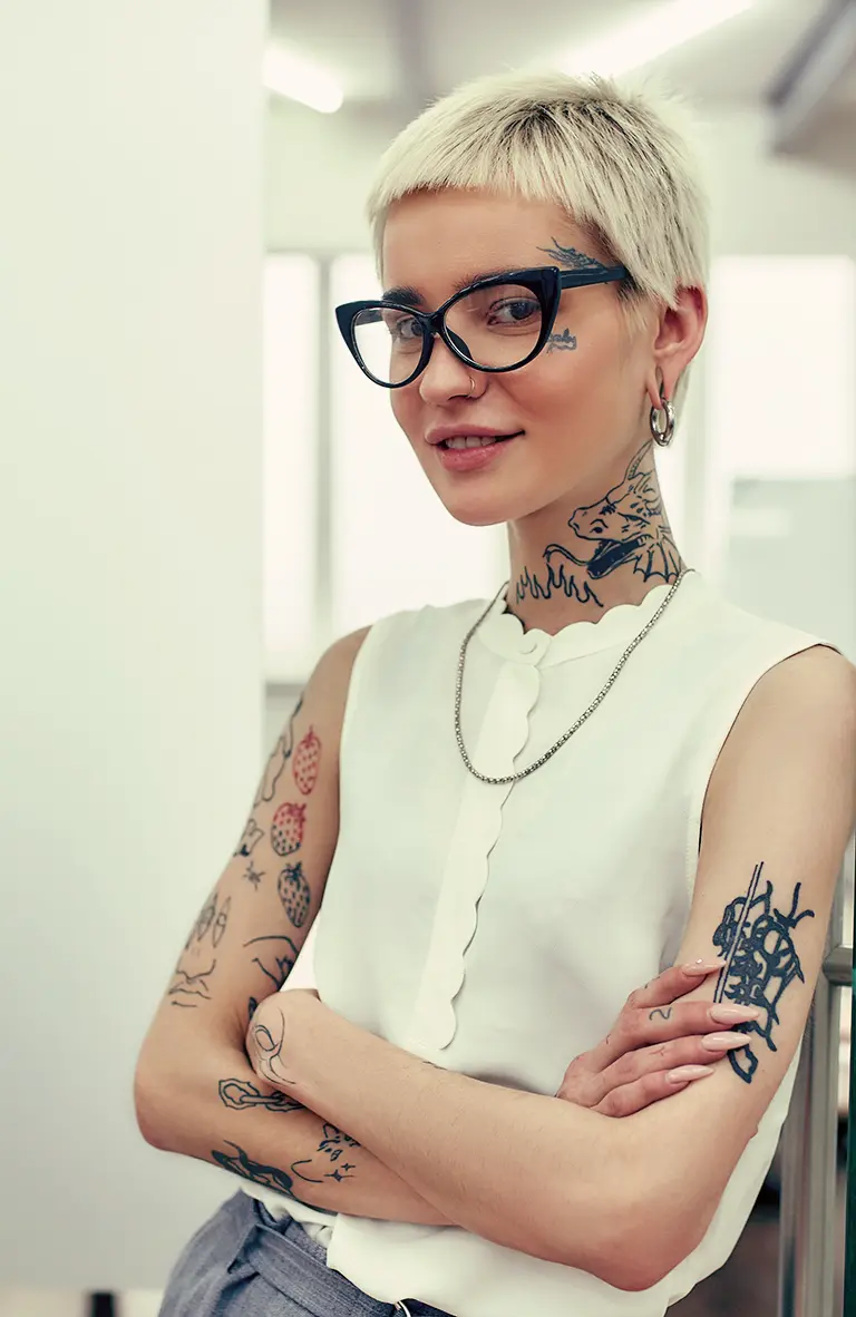 A young woman with short hair and tattoos leans against a doorframe and looks directly at ou as if listening intently